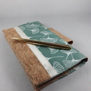 May include: A journal with a cork cover and a teal fabric top featuring a white leaf pattern. A gold pen rests on the journal. A white ribbon accents the journal's spine. The journal is a stationery item.