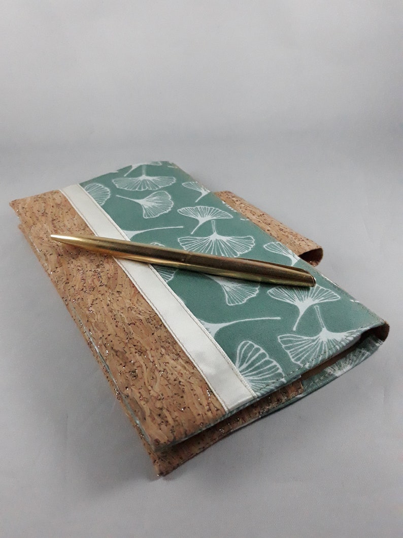 May include: A journal with a cork cover and a teal fabric top featuring a white leaf pattern. A gold pen rests on the journal. A white ribbon accents the journal's spine. The journal is a stationery item.