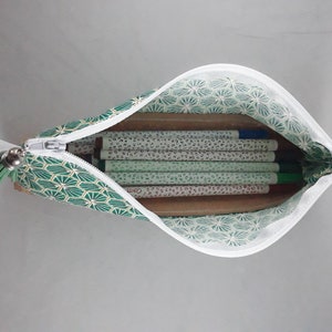 May include: Open pencil case with a white zipper, filled with pens and pencils. The case is green with a geometric pattern and a white interior. A small green tassel hangs from the zipper.