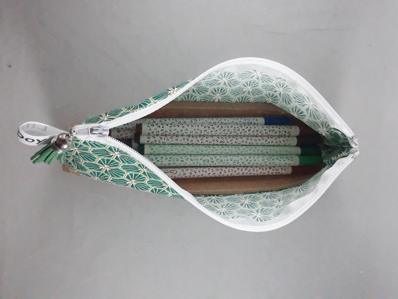 May include: Open pencil case with a white zipper, filled with pens and pencils. The case is green with a geometric pattern and a white interior. A small green tassel hangs from the zipper.