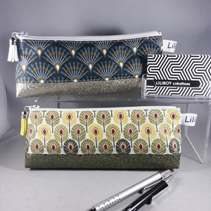 May include: Two pencil cases with zipper closures. One is navy blue with a fan-like pattern and a silver glitter base. The other is white with a leaf pattern and a green glitter base. The cases have white and yellow tassels. A business card with the text "LILIBOY cr&eacute;ations" is also visible.