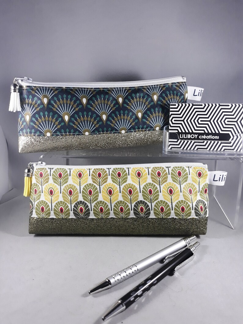 May include: Two pencil cases with zipper closures. One is navy blue with a fan-like pattern and a silver glitter base. The other is white with a leaf pattern and a green glitter base. The cases have white and yellow tassels. A business card with the text "LILIBOY cr&eacute;ations" is also visible.