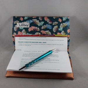 May include: A checkbook cover with a teal and floral pattern, the word "Liliboy" is printed on the cover. Inside, a teal pen rests on a check with printed text. The cover is open, revealing a brown leather-like interior.