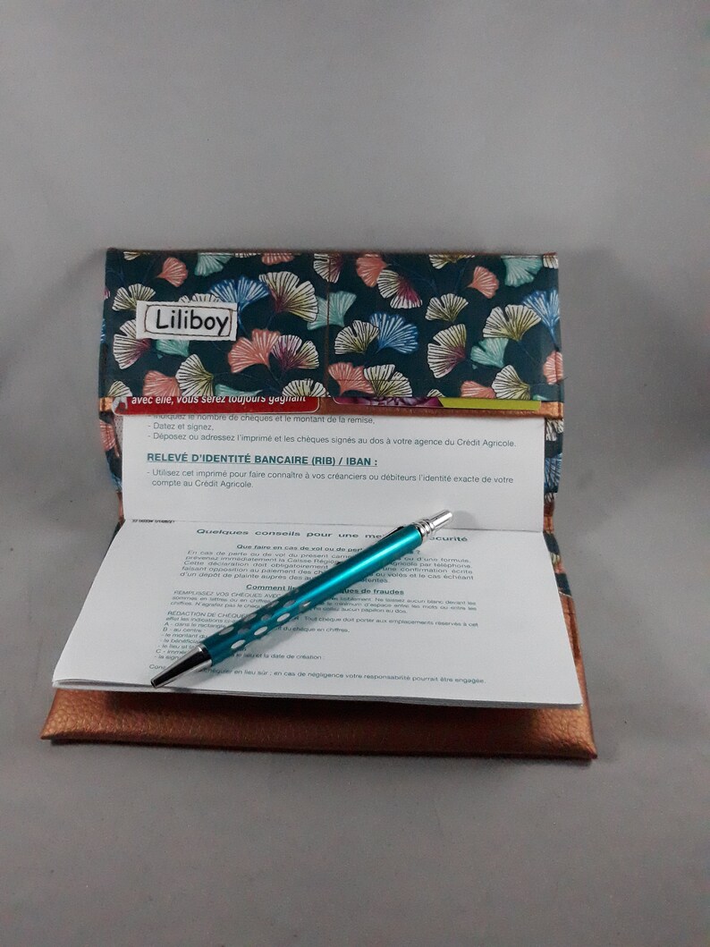 May include: A checkbook cover with a teal and floral pattern, the word "Liliboy" is printed on the cover. Inside, a teal pen rests on a check with printed text. The cover is open, revealing a brown leather-like interior.