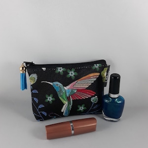 Op de afbeelding: Een zwarte cosmetische tas met een kleurrijk kolibrie-ontwerp, een blauwe kwast, een lippenstift en een fles blauwe nagellak.