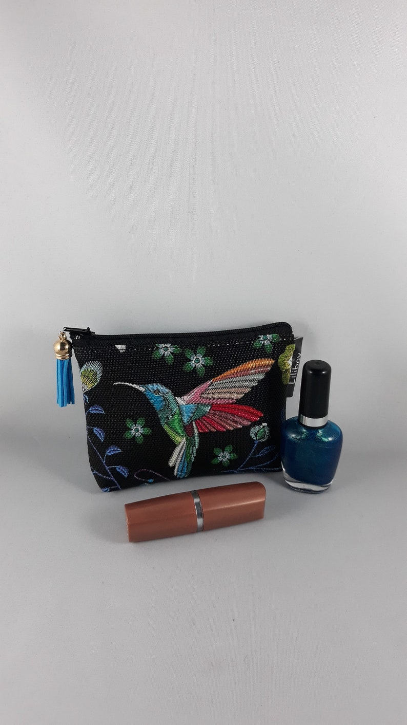 Op de afbeelding: Een zwarte cosmetische tas met een kleurrijk kolibrie-ontwerp, een blauwe kwast, een lippenstift en een fles blauwe nagellak.