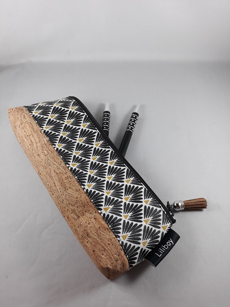 May include: A pencil case with a black and white floral pattern and a cork base. The pencil case has a black zip and a brown tassel. Two black and white pens are inside the case. The label says "Liliboy".