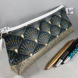 May include: A rectangular pencil case with a black background and a fan-like pattern in teal, white, and gold. The bottom edge has a gold glitter trim. A white zipper and tassel complete the design. The label reads "Lili".