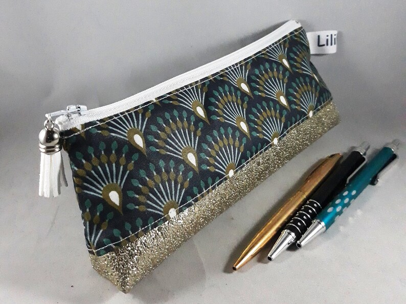 May include: A rectangular pencil case with a black background and a fan-like pattern in teal, white, and gold. The bottom edge has a gold glitter trim. A white zipper and tassel complete the design. The label reads "Lili".