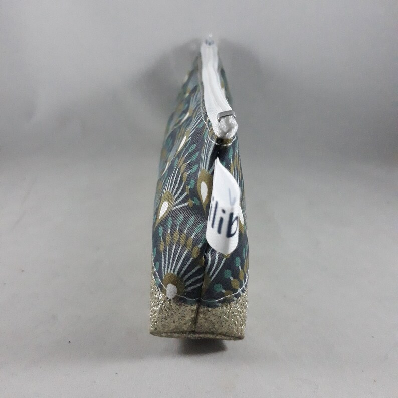 May include: A small, rectangular cosmetic bag with a white zipper and a gold glitter base. The bag features a dark gray floral pattern with teal and gold accents. A white tag with the word "lib" is attached.