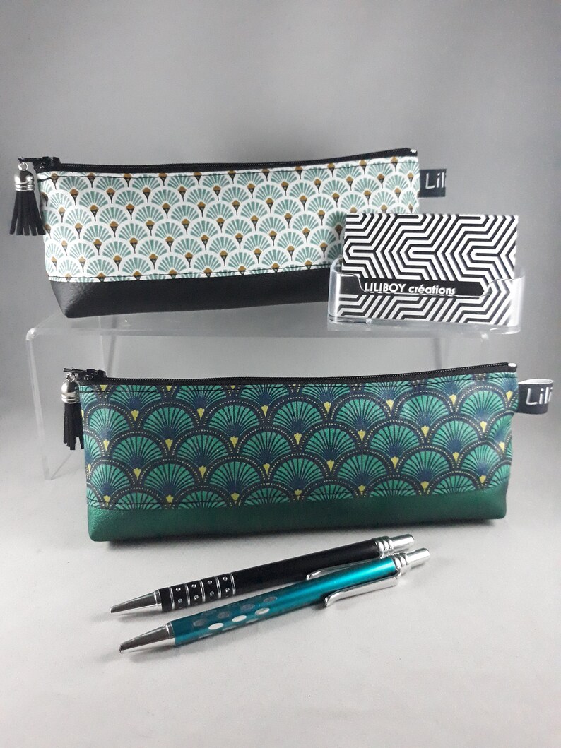 May include: Two pencil cases with a fan-shaped pattern in teal, gold, and white, with black trim and zippers. One case has a black tassel. The image also shows two pens and a business card holder with the text "LILIBOY cr&eacute;ations".
