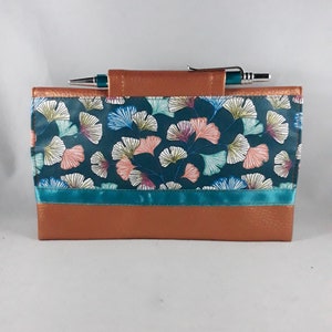 May include: A teal and brown journal cover with a pen. The cover features a teal background with a floral pattern in pink, white, and blue. A teal ribbon accents the design. The top and bottom are brown.