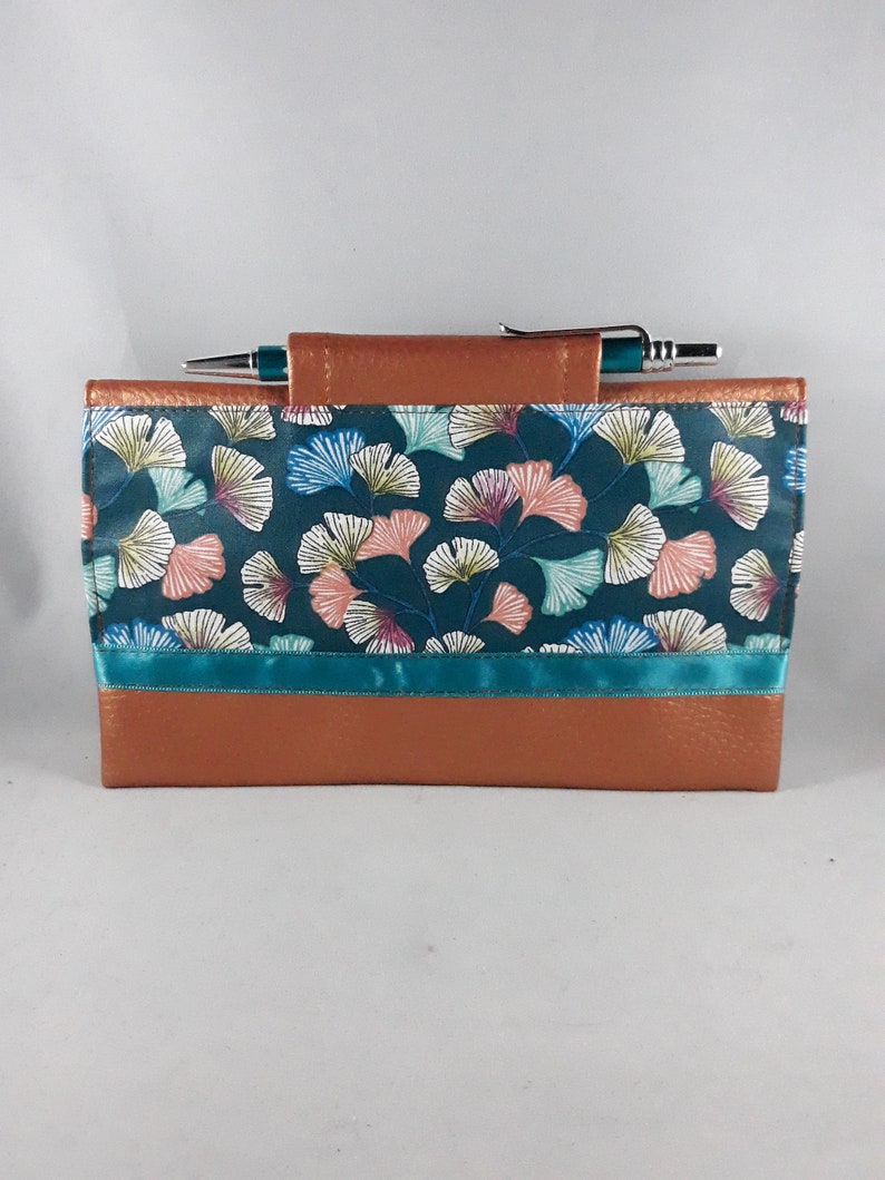 May include: A teal and brown journal cover with a pen. The cover features a teal background with a floral pattern in pink, white, and blue. A teal ribbon accents the design. The top and bottom are brown.