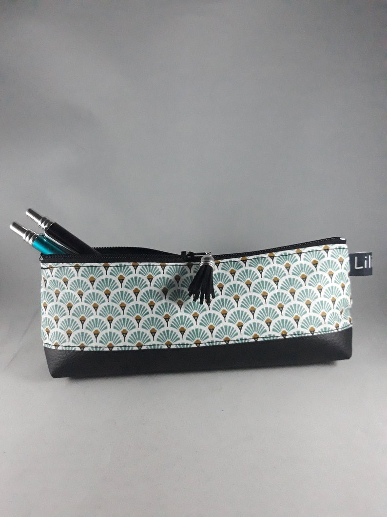 May include: A rectangular pencil case with a black zipper and tassel. The case features a pattern of teal and gold fan shapes on a white background, with a black faux leather base. Two pens are visible inside.