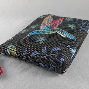 Op de afbeelding: Zwarte etui met kleurrijke borduursels van een kolibrie en bloemen. De kolibrie heeft blauwe, groene en rode vleugels. Een roze kwastje hangt aan de ritssluiting. Rechthoekige vorm.