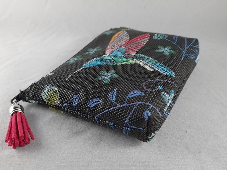 Op de afbeelding: Zwarte etui met kleurrijke borduursels van een kolibrie en bloemen. De kolibrie heeft blauwe, groene en rode vleugels. Een roze kwastje hangt aan de ritssluiting. Rechthoekige vorm.
