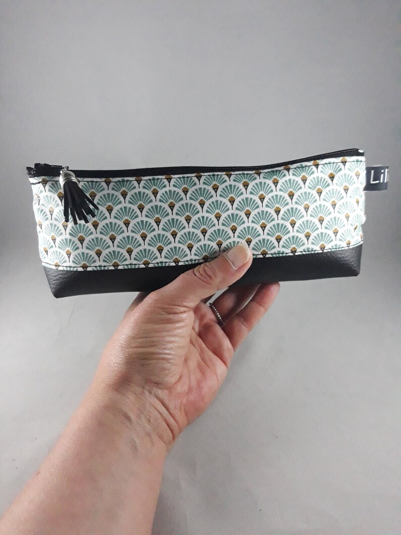 May include: A rectangular pencil case with a black zipper and tassel. The top portion features a white fabric with a teal and gold fan pattern. The bottom is black. The word "Lil" is printed on a small black tag.
