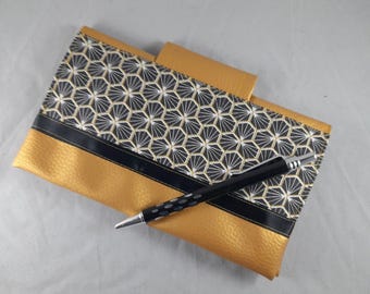 May include: A rectangular gold and black wallet featuring a geometric honeycomb pattern on the upper section, a solid gold lower section, and a thin black horizontal stripe. A black ballpoint pen with silver accents rests diagonally across the wallet.