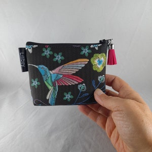 Op de afbeelding: Een kleine, zwarte etui met ritssluiting met een kleurrijk kolibrie-ontwerp en bloemaccenten. De kolibrie heeft blauwe, groene en roze veren. Een roze kwastje hangt aan de ritstrekker. De merknaam "Liliboy" staat op een klein zwart label.