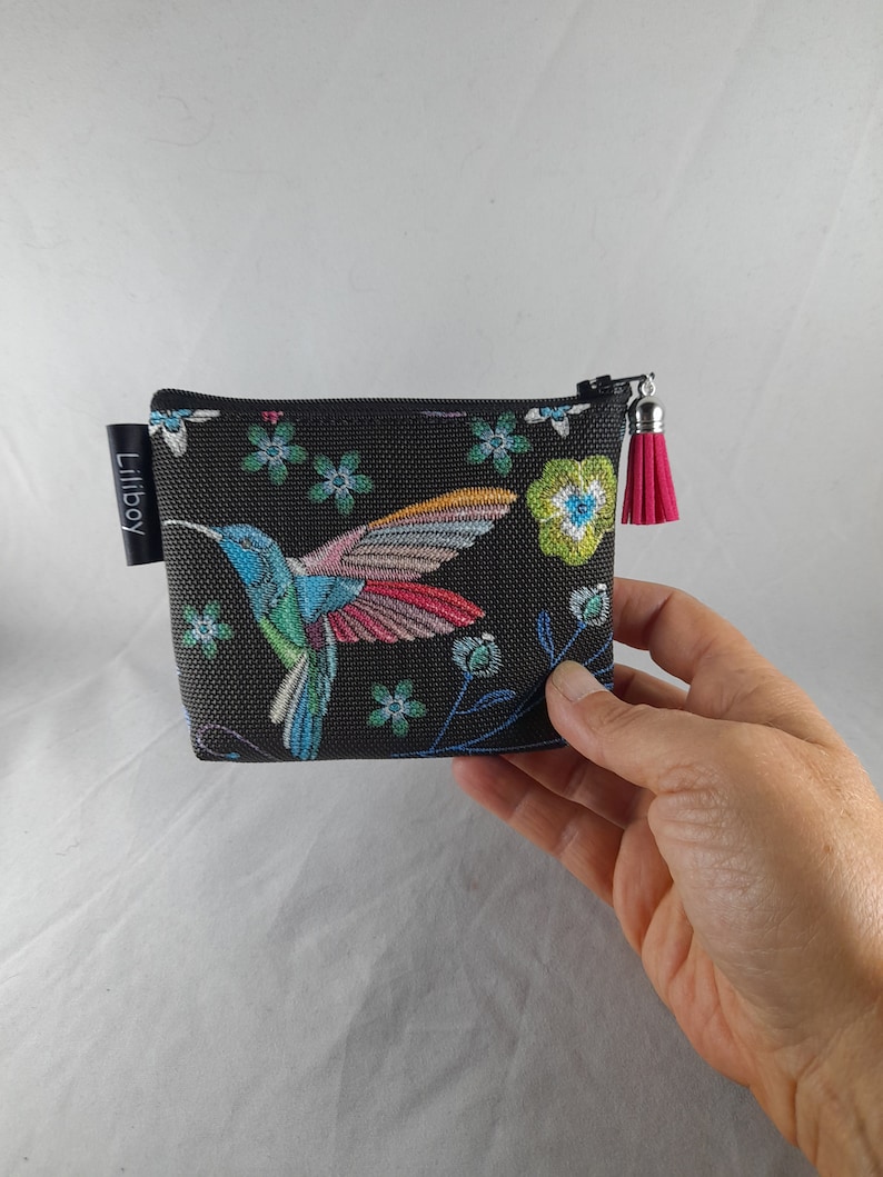 Op de afbeelding: Een kleine, zwarte etui met ritssluiting met een kleurrijk kolibrie-ontwerp en bloemaccenten. De kolibrie heeft blauwe, groene en roze veren. Een roze kwastje hangt aan de ritstrekker. De merknaam "Liliboy" staat op een klein zwart label.