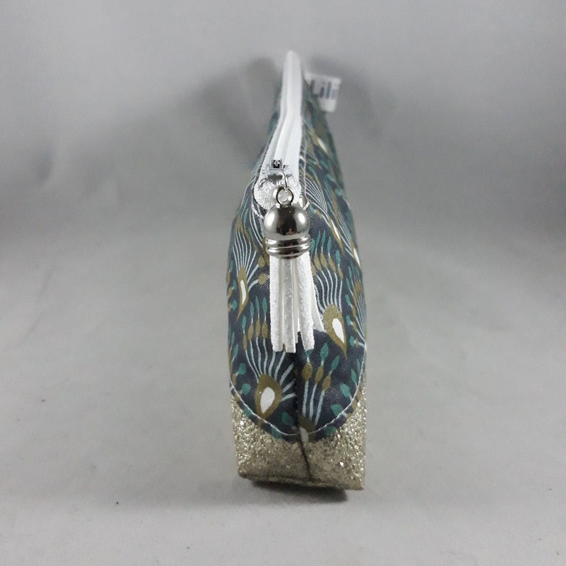 May include: A small, zippered cosmetic bag with a patterned fabric in teal, gold, and white. The bottom of the bag is a gold glitter material. The zipper pull has a white tassel.