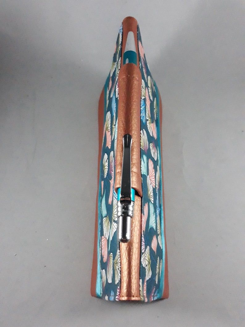May include: A pen case with a teal and copper color scheme. The case features a pattern of stylized leaves in shades of pink, blue, and yellow. The pen case is made of a textured material and has a copper-colored zipper and trim.