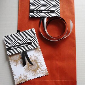 May include: An orange paper gift bag with two tags and a ribbon. The tags have a black and white geometric pattern and the text "LILIBOY cr&eacute;ations". One tag features a sunflower design. A black and silver ribbon is curled on the bag.