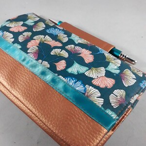 May include: A handmade journal with a teal and copper color scheme. The top portion features a teal fabric with a floral pattern, accented by a teal ribbon. The bottom is copper-colored leather. A pen is tucked into a leather pen holder.
