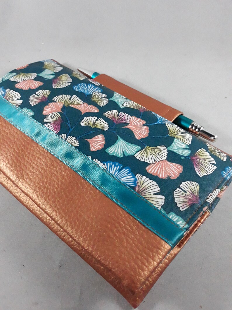 May include: A handmade journal with a teal and copper color scheme. The top portion features a teal fabric with a floral pattern, accented by a teal ribbon. The bottom is copper-colored leather. A pen is tucked into a leather pen holder.
