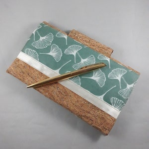 May include: A cork notebook with a teal cover featuring a white ginkgo leaf pattern. A gold pen rests on the notebook. A white ribbon accents the teal cover. The notebook is a natural color with a textured surface.