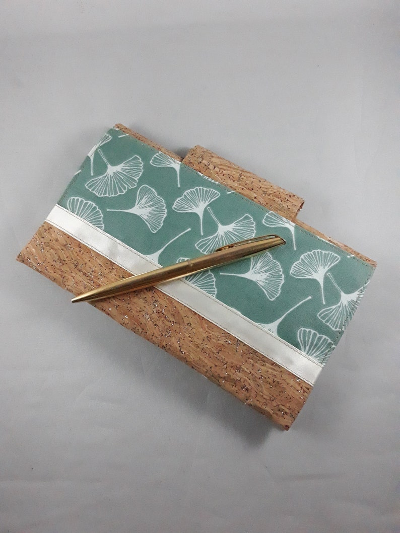 May include: A cork notebook with a teal cover featuring a white ginkgo leaf pattern. A gold pen rests on the notebook. A white ribbon accents the teal cover. The notebook is a natural color with a textured surface.