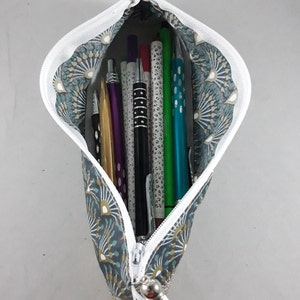 May include: A fabric pencil case with a white zipper, filled with various pens and pencils. The case has a teal and gray pattern with a white tassel. The pencils include gold, purple, black, green, and blue.