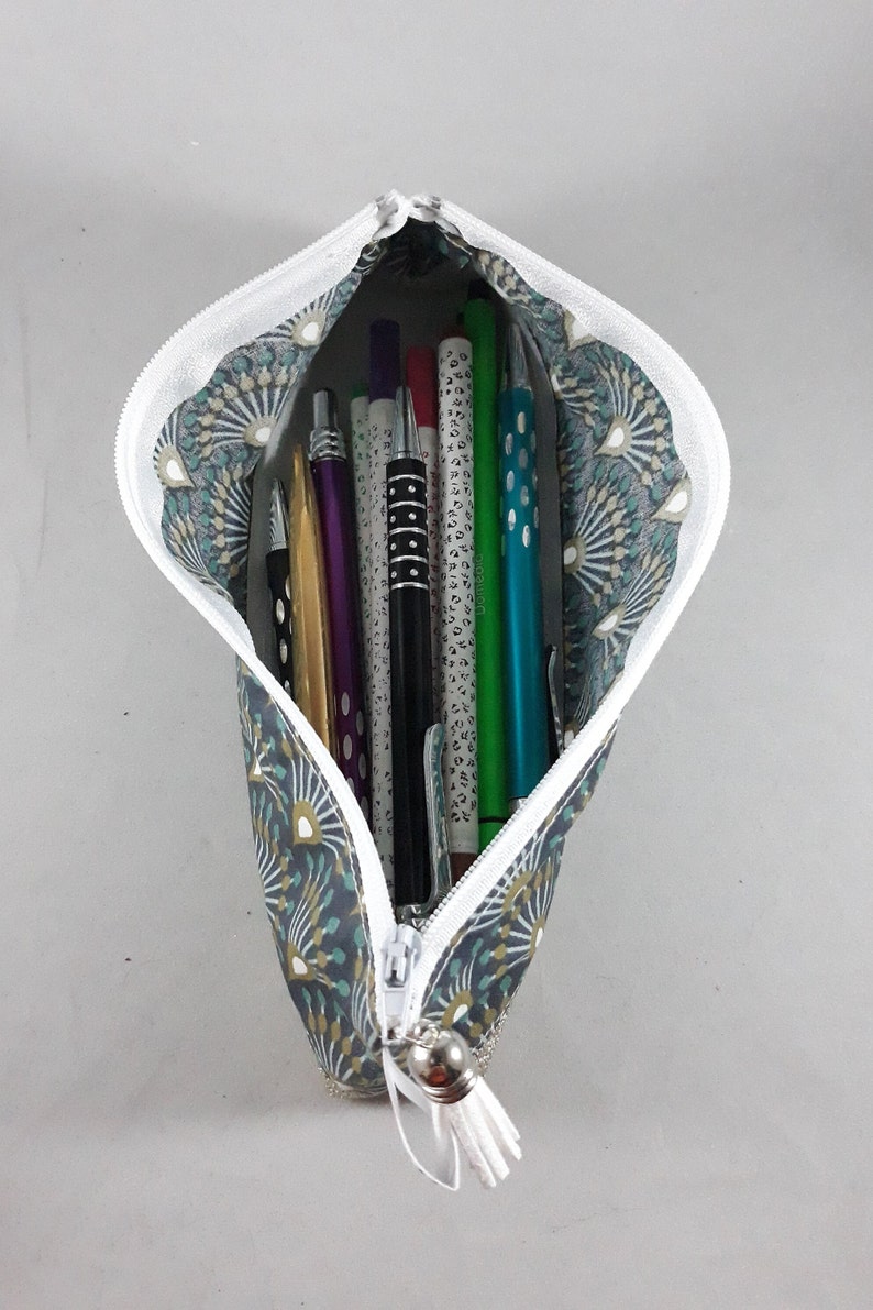 May include: A fabric pencil case with a white zipper, filled with various pens and pencils. The case has a teal and gray pattern with a white tassel. The pencils include gold, purple, black, green, and blue.
