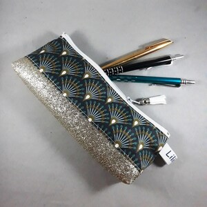May include: A rectangular pencil case with a white zipper and a tassel. The case features a dark teal fan pattern and a gold glitter accent. Three pens are partially visible inside the case.