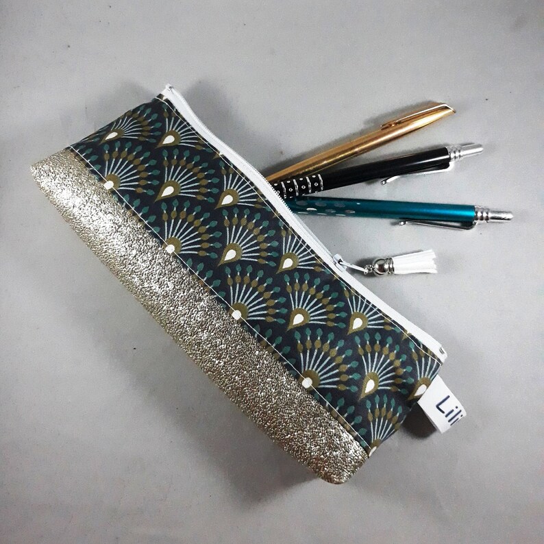 May include: A rectangular pencil case with a white zipper and a tassel. The case features a dark teal fan pattern and a gold glitter accent. Three pens are partially visible inside the case.