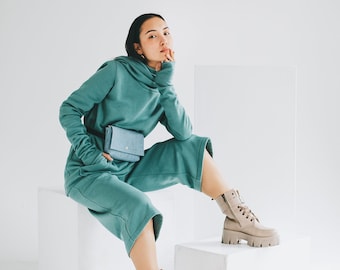 May include: A woman wearing a teal green hooded sweatshirt and matching pants with a small blue bag worn as a belt. She is sitting on a white cube with her legs crossed and wearing beige ankle boots.