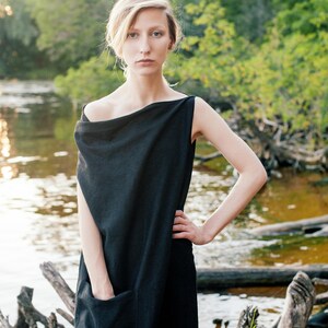 May include: A woman wearing a black, sleeveless, draped dress with a single pocket. She is standing in front of a body of water with a natural background.