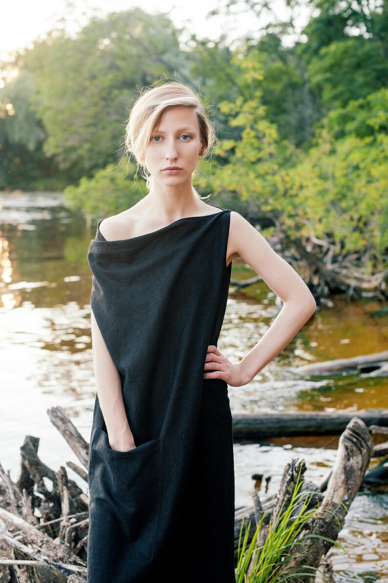May include: A woman wearing a black, sleeveless, draped dress with a single pocket. She is standing in front of a body of water with a natural background.