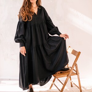 May include: A woman in a long, black dress with a tiered skirt and long sleeves. The dress has a V-neck and is worn with black shoes. She is standing beside a wooden chair.