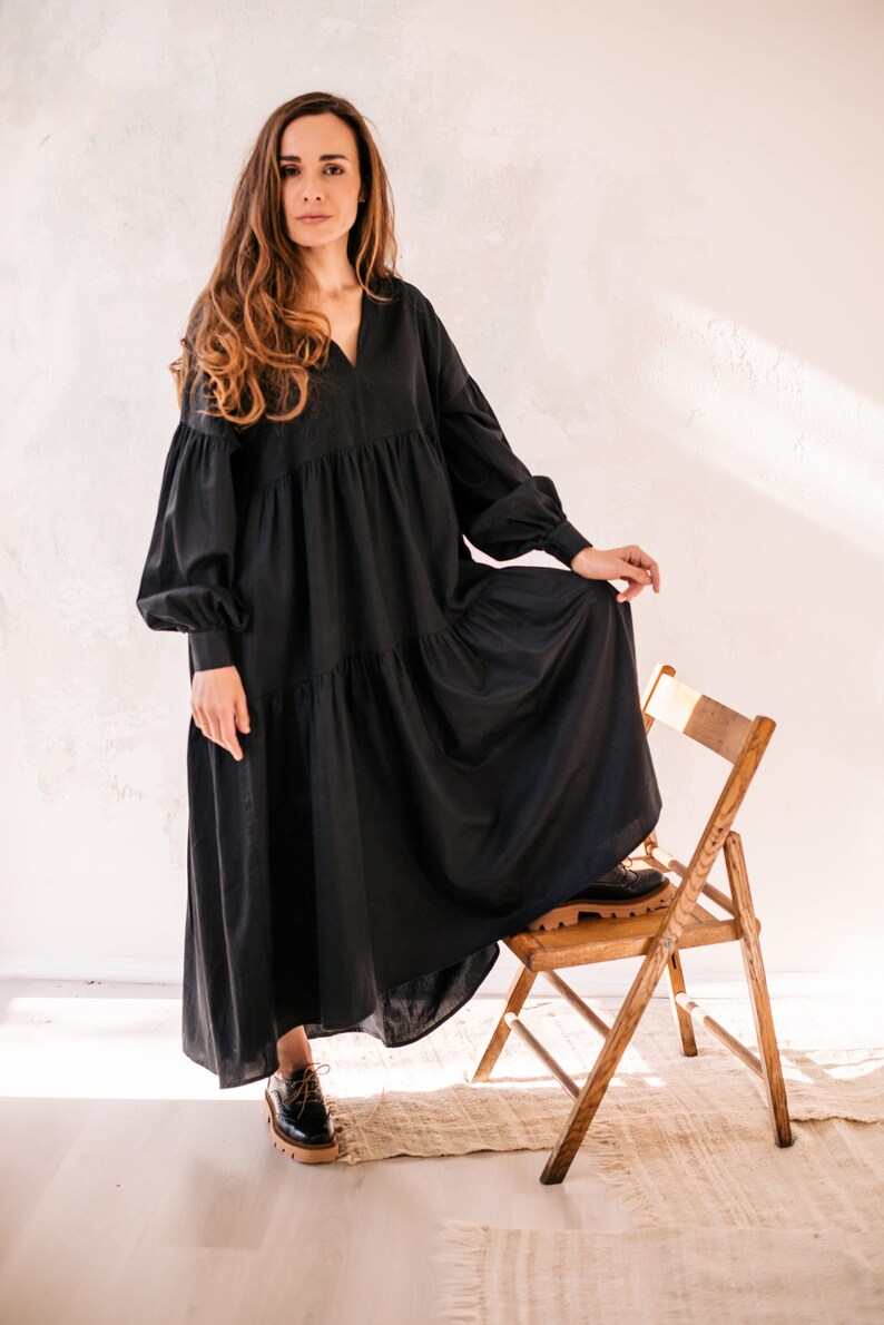 May include: A woman in a long, black dress with a tiered skirt and long sleeves. The dress has a V-neck and is worn with black shoes. She is standing beside a wooden chair.