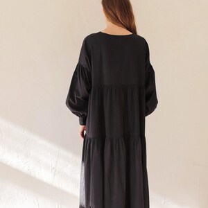 May include: A black dress featuring a tiered skirt and long, voluminous sleeves. The dress has a round neckline and a relaxed fit. The model is wearing grey shoes. The dress is made of a soft, flowing fabric.