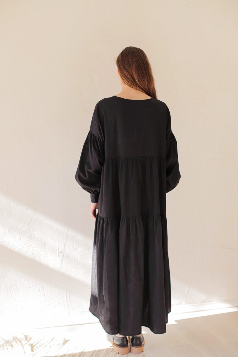 May include: A black dress featuring a tiered skirt and long, voluminous sleeves. The dress has a round neckline and a relaxed fit. The model is wearing grey shoes. The dress is made of a soft, flowing fabric.