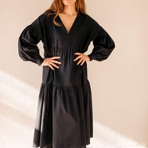 May include: A black midi dress with a V-neck and long, voluminous sleeves. The dress has a tiered skirt and is worn with black leather boots with tan soles and yellow laces. The model is standing in front of a cream-coloured wall.