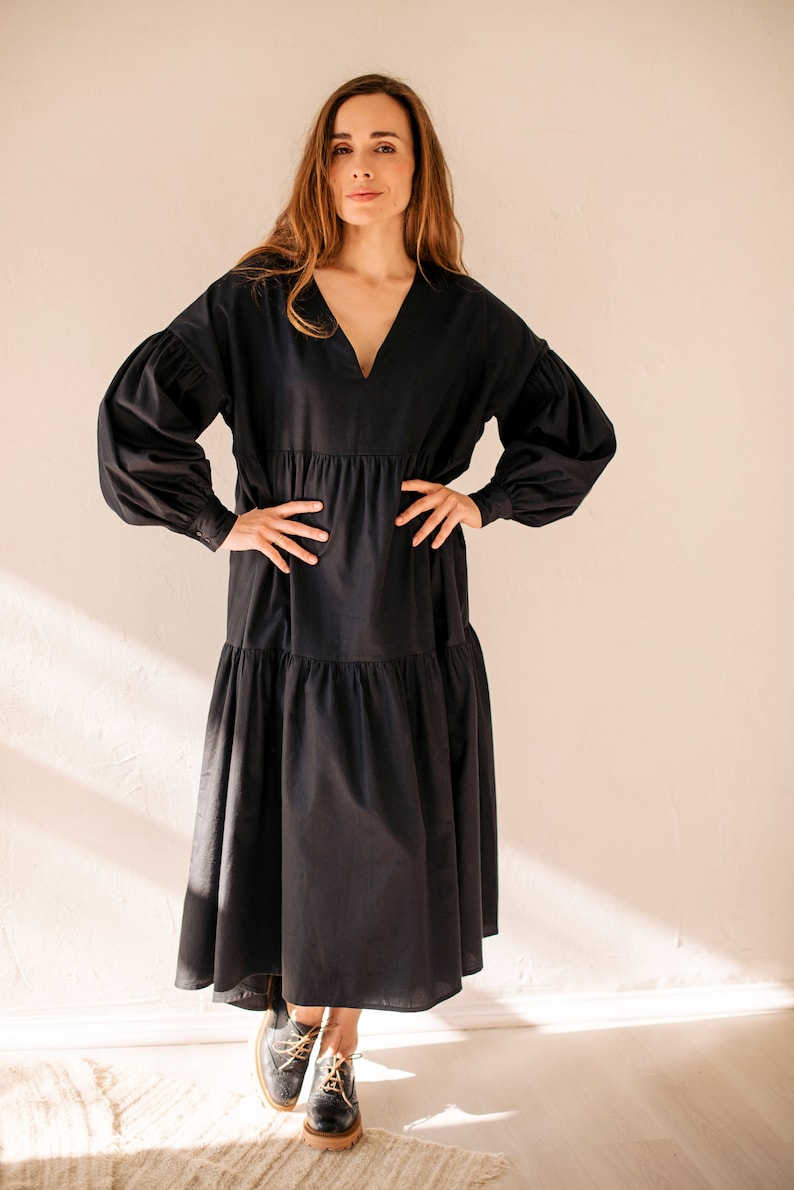 May include: A black midi dress with a V-neck and long, voluminous sleeves. The dress has a tiered skirt and is worn with black leather boots with tan soles and yellow laces. The model is standing in front of a cream-coloured wall.