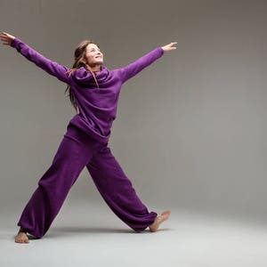 May include: A woman wearing a purple jumpsuit with a cowl neck, standing with her arms outstretched and looking up.