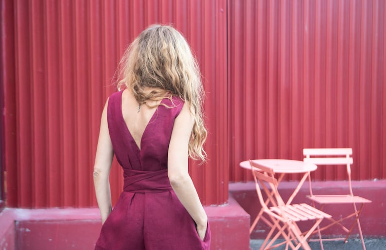 May include: A woman wearing a burgundy sleeveless jumpsuit with a deep V-back and a tie at the waist. She is standing in front of a red corrugated metal wall with two pink metal chairs and a table in the foreground.