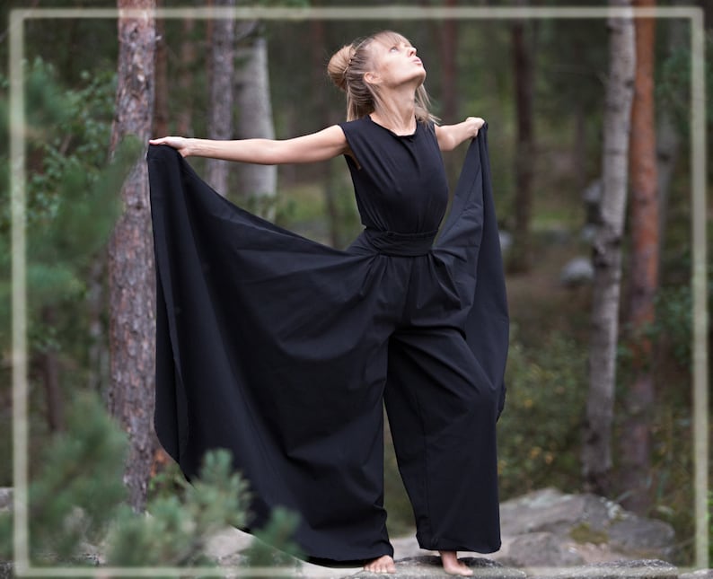 May include: A woman wearing a black jumpsuit with wide legs and a flowing skirt-like design. She is standing in a forest with trees in the background.