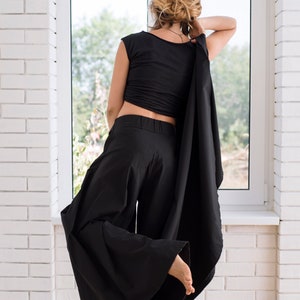 May include: A woman wearing a black top and wide-legged pants with a long black scarf draped over her shoulders. She is standing in front of a window with her back to the camera.