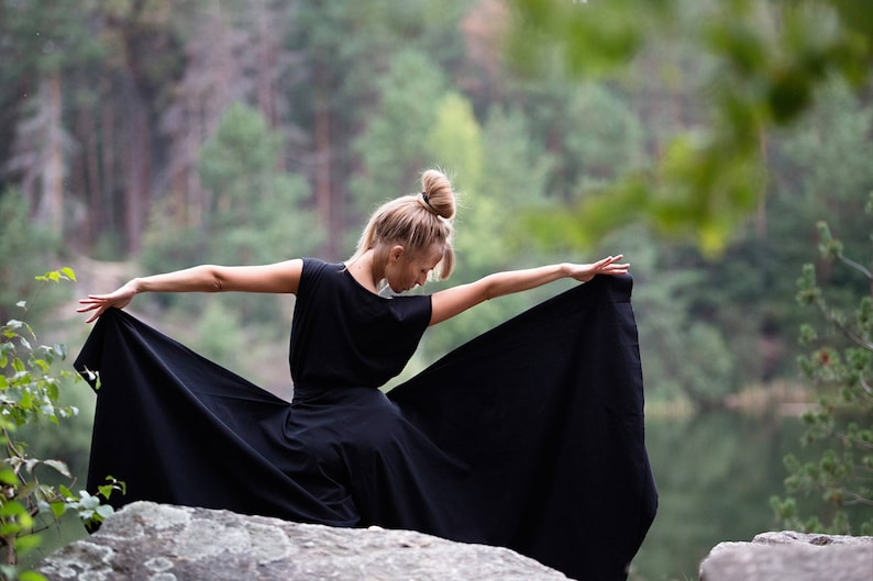 May include: A woman in a black dress stands on a rock with a forest background. She is holding the dress out to the sides, creating a flowing silhouette.