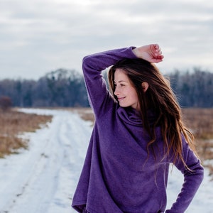 May include: A woman wearing a purple long-sleeved top and matching pants stands in a snowy field. She is looking off to the side and has her hand on her head.